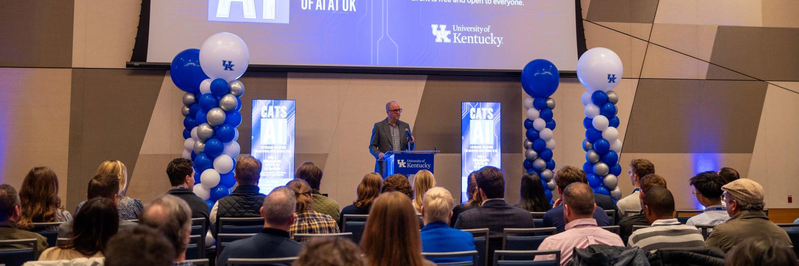 man speaking at podium to large room in front of large screen displaying CATS AI and Microsoft AI In Action event