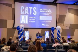 man speaking at podium to large room in front of large screen displaying CATS AI and Microsoft AI In Action event