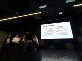 Man moderating a panel discussion with three others sitting on stage in front of large presentation screen