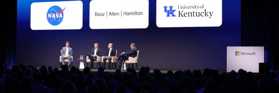 four men sitting on a stage to talk on a panel in front of audience